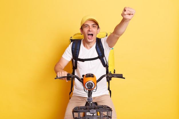 Porträt eines positiven, glücklichen, optimistischen Lieferboten auf dem Fahrrad mit weißem T-Shirt und Kappe isoliert auf gelbem Hintergrund, erhobene Faust, die Ankündigung macht