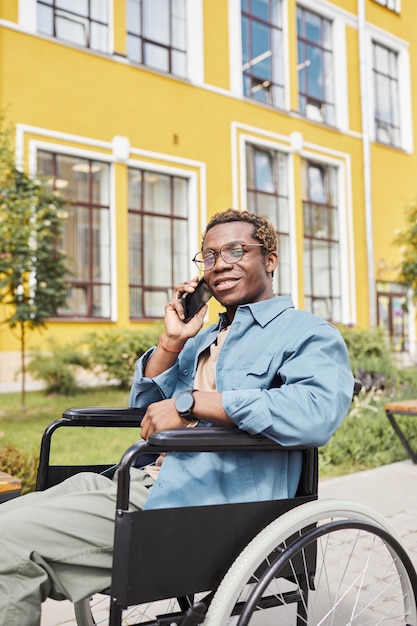 Porträt eines positiven gutaussehenden jungen behinderten afroamerikanischen Mannes mit Brille, der im Rollstuhl sitzt und telefonisch anruft