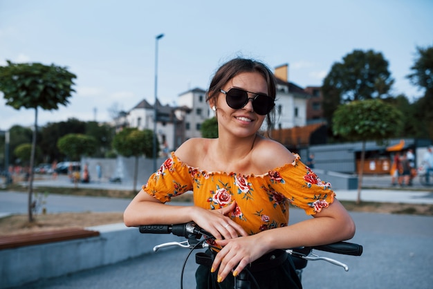 Porträt eines positiven jungen Mädchens mit Sonnenbrille, das mit ihrem Fahrrad steht.