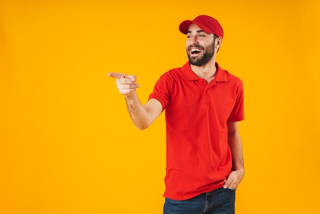 Porträt eines positiven Lieferboten in rotem T-Shirt und Mütze, der lächelt und mit dem Finger auf das Exemplar isoliert über Gelb zeigt