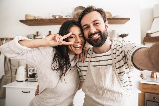 Porträt eines positiven Paares, der Schürzen trägt und ein Selfie-Foto beim Kochen in der Küche zu Hause macht