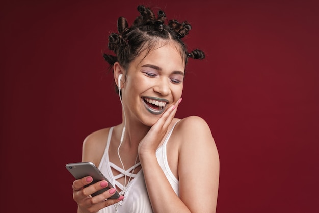 Porträt eines positiven Punk-Mädchens mit bizarrer Frisur und dunklem Lippenstift, das lacht, während es Smartphone mit Kopfhörern verwendet, die über roter Wand isoliert sind?