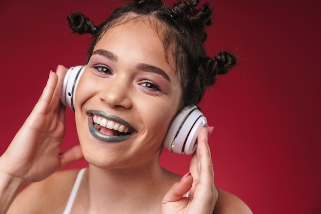 Porträt eines positiven Punk-Mädchens mit bizarrer Frisur und dunklem Lippenstift, das lächelt, während sie Musik mit Kopfhörern hört, die über roter Wand isoliert sind?