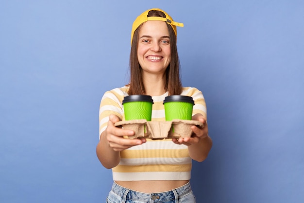 Porträt eines positiven Teenager-Mädchens mit gestreiftem T-Shirt und Baseballkappe, das isoliert vor blauem Hintergrund posiert. Frau, die als Kurier arbeitet und Kaffee zum Mitnehmen liefert