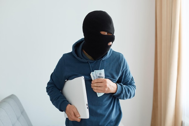 Porträt eines Räubers mit blauem Hoodie und schwarzer Sturmhaube, der Laptop und Bargeld im Schlauch einer Person ausraubt, den Ort des Verbrechens verlässt, das Fenster ansieht, illegale Handlungen.