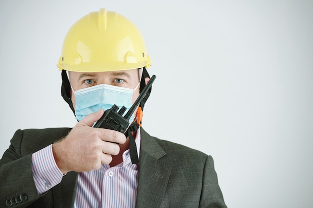 Porträt eines reifen Bauunternehmers in gelbem Bauarbeiterhelm und Maske mit Walkie-Talkie während der Übertragung von Nachrichten