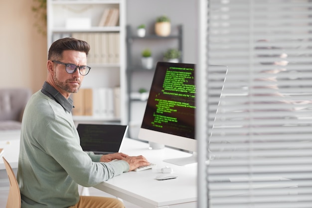 Porträt eines reifen Entwicklers, der nach vorne schaut, während er am Tisch mit dem Computer in der Softwareentwicklungsfirma arbeitet