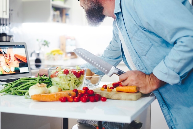 Porträt eines reifen männlichen Mannes zu Hause, der ein gesundes, leckeres Frühstück nach Rezept auf einem digitalen Tablet zubereitet, während er mit Hilfe eines kulinarischen Online-Tutorials in der heimischen Küche kocht Ehemann kocht