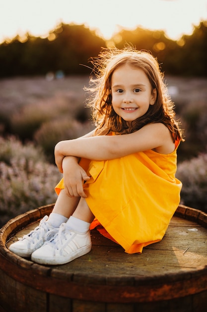 Porträt eines reizenden kleinen Mädchens, das auf einem Fass sitzt und Kamera lachend betrachtet, während sie ihre Hand auf ihren Beinen gegen Sonnenuntergang in einem Lavendelfeld reinigt.