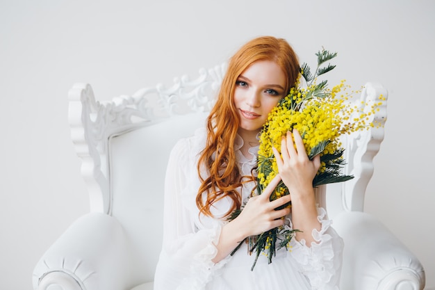 Porträt eines rothaarigen schönen Mädchens mit einer Mimose in einem langen weißen Kleid