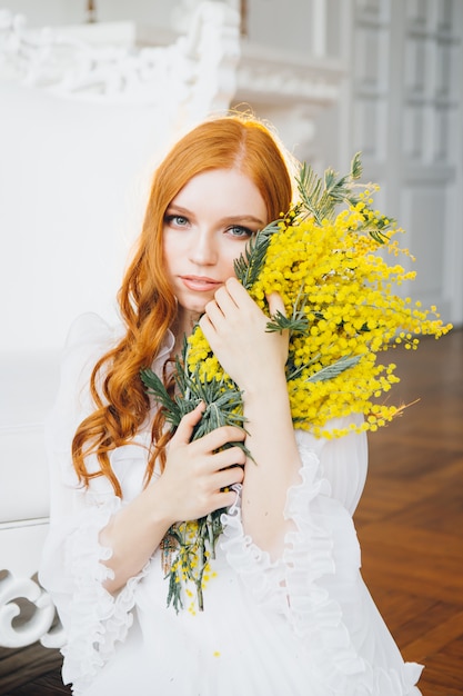 Porträt eines rothaarigen schönen Mädchens mit einer Mimose in einem langen weißen Kleid