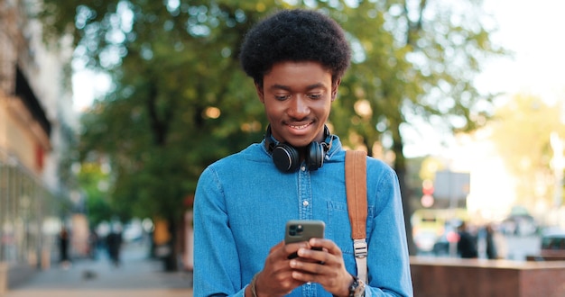 Porträt eines ruhigen, brünetten, gemischtrassigen Mannes, der legere Kleidung trägt und auf den Smartphone-Bildschirm schaut, während er am Frühlingstag auf der grünen Straße steht und seine Zähne beim breiten Lächeln zeigt