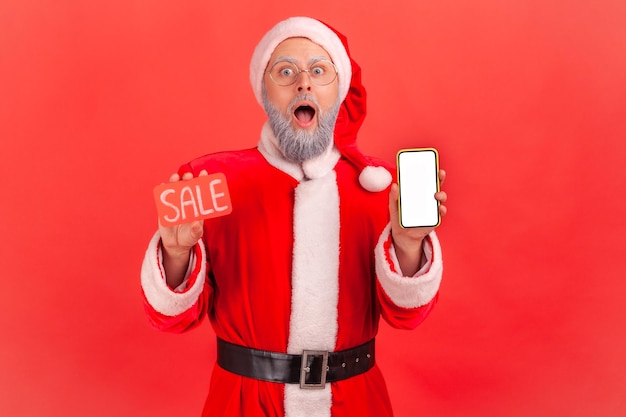 Porträt eines schockierten älteren Mannes mit grauem Bart im Weihnachtsmannkostüm, der sein Smartphone in der Hand hält