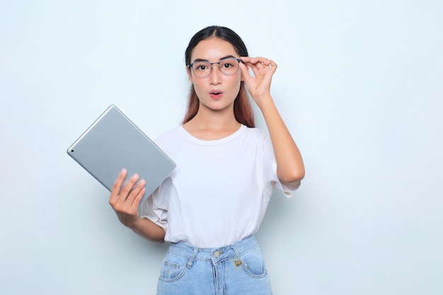 Porträt eines schockierten jungen asiatischen Mädchens im weißen T-Shirt, das eine digitale Tablette hält, die eine Brille isoliert auf weißem Hintergrund berührt