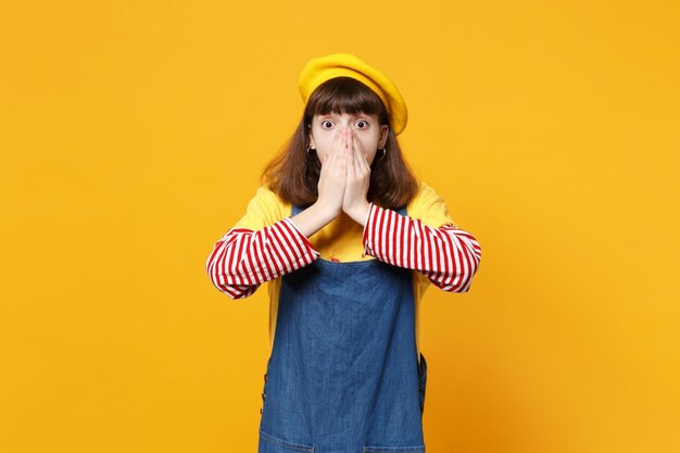 Porträt eines schockierten Mädchen-Teenagers in französischer Baskenmütze, Denim-Sommerkleid, das den Mund mit den Händen bedeckt, isoliert auf gelbem Wandhintergrund im Studio. Menschen aufrichtige Emotionen Lifestyle-Konzept. Mock-up-Kopienbereich.