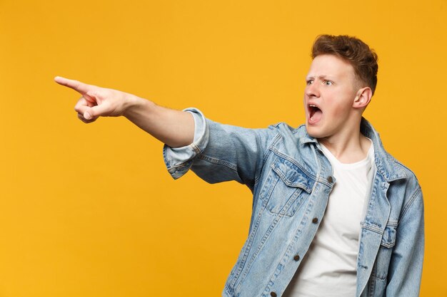 Porträt eines schockierten, schreienden jungen Mannes in Denim-Freizeitkleidung, der den Zeigefinger zur Seite zeigt, isoliert auf gelb-orangefarbenem Wandhintergrund. Menschen aufrichtige Emotionen, Lifestyle-Konzept. Mock-up-Kopienbereich.