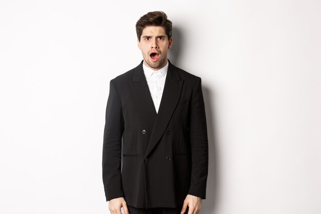Porträt eines schockierten und erschrockenen gutaussehenden Mannes in Anzug, Kiefer und Blick in Ehrfurcht in die Kamera, stehend auf weißem Hintergrund