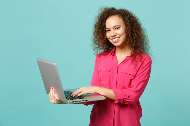 Porträt eines schönen afrikanischen Mädchens in rosa Freizeitkleidung mit Laptop-PC isoliert auf blauem türkisfarbenem Wandhintergrund im Studio. Menschen aufrichtige Emotionen, Lifestyle-Konzept. Kopieren Sie Platz.