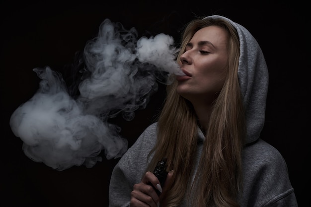 Porträt eines schönen blonden Mädchens in grauem Hoodie raucht Vape einzeln auf schwarzem Studiohintergrund, Dampfrauchwolke, Mini-Shisha