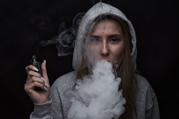 Porträt eines schönen blonden Mädchens in grauem Hoodie raucht Vape einzeln auf schwarzem Studiohintergrund, Dampfrauchwolke, Mini-Shisha