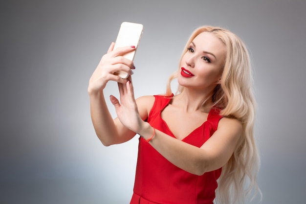 Porträt eines schönen erfolgreichen lächelnden Mädchens, das selfie im roten Kleid auf hellem Hintergrund macht