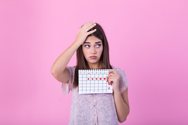 Porträt eines schönen jungen Mädchens, das ihren Menstruationskalender hält und ihr Gesicht über einem rosa Hintergrund abdeckt.