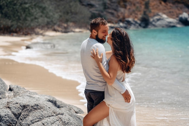 Porträt eines schönen jungen Paares, das an einem Sandstrand neben einem tropisch glänzenden Meer posiert und sich mit liebevollen Augen ansieht.