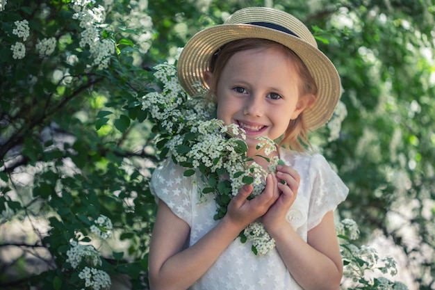 Porträt eines schönen kaukasischen kleinen Mädchens in Boho-Kleid und einem Hut im blühenden Busch mit weißen Spirea-Blumen mit selektivem Fokus und weichem Fokus.