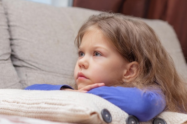 Porträt eines schönen kaukasischen kleinen Mädchens von 5 Jahren, das auf der Couch liegt, den Kopf auf die Hände legt und sehr aufmerksam drinnen zur Seite schaut
