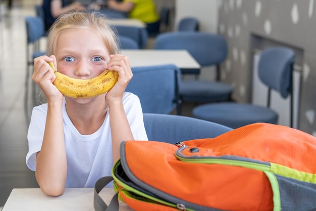 Porträt eines schönen kleinen Mädchens mit einem Bananenlächeln, das mit einem Schulrucksack an einem Tisch im Klassenzimmer sitzt Gesundes Essen in der Schule