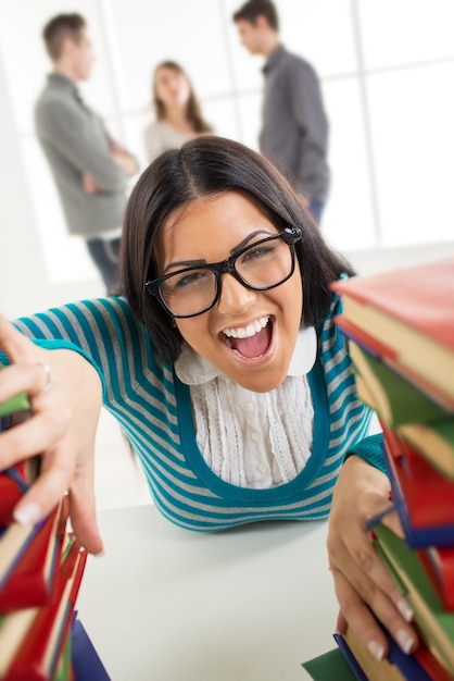 Porträt eines schönen lustigen Studentenmädchens, das unter den vielen Büchern im Vordergrund sitzt und schreit. Eine ihrer Freunde steht hinter ihr und redet. Blick in die Kamera.