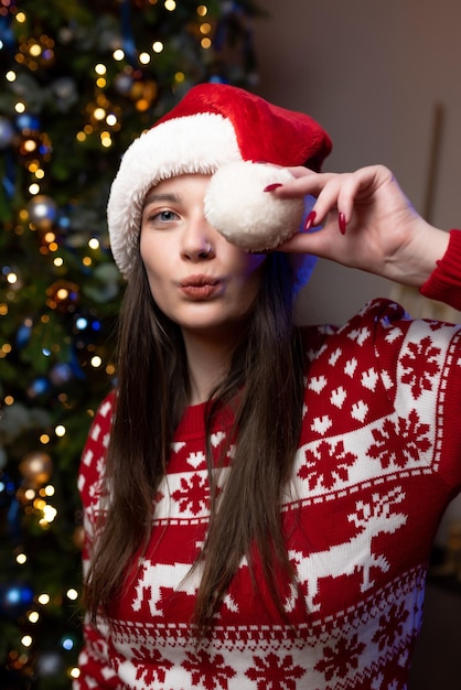 Porträt eines schönen Mädchens in einem Weihnachtspullover und einem roten Weihnachtsmann-Hut sitzt in der Nähe eines luxuriösen Weihnachtsbaums. Posiert und lächelnd. Feier des neuen Jahres, Weihnachten