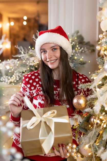 Porträt eines schönen Mädchens in einem Weihnachtspullover und einem roten Weihnachtsmann-Hut steht in der Nähe eines luxuriösen Weihnachtsbaums. Lächelnd und mit einem Geschenk in den Händen. Feier des neuen Jahres, Weihnachten