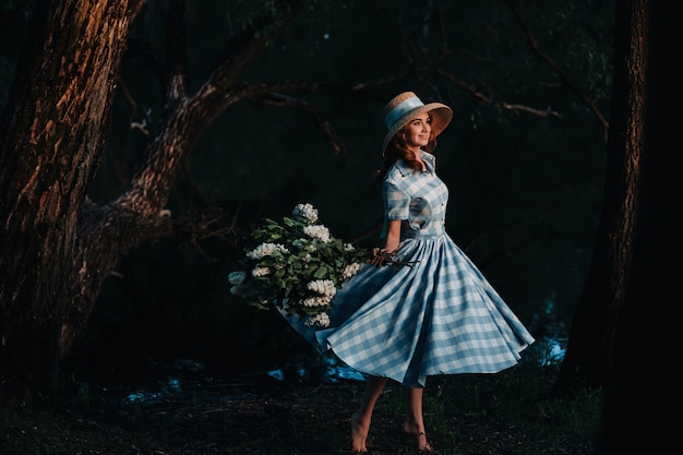 Porträt eines schönen Mädchens mit langen Haaren, einem Strohhut und einem langen Sommerkleid mit lila Blumen im Garten