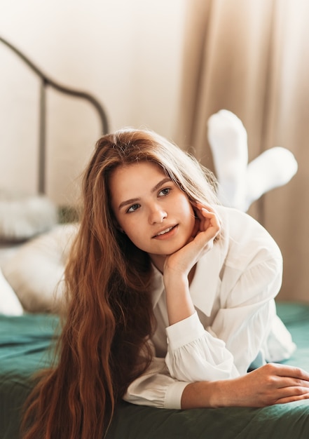 Porträt eines schönen Mädchens mit langen Haaren in weißer Kleidung im Schlafzimmer. Eine junge Frau ruht auf einem Bett