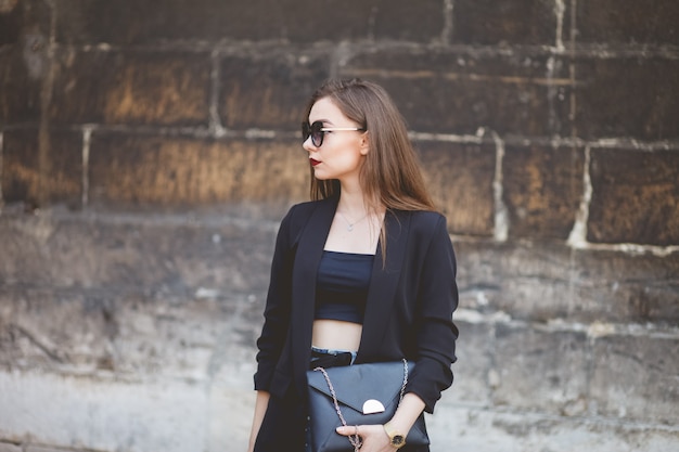 Porträt eines schönen Mädchens mit Sonnenbrille und eine Tasche in seinen Händen auf dem Hintergrund einer Steinmauer