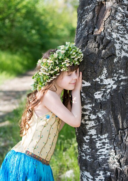 Porträt eines schönen Mädchens von neun Jahren mit langen braunen Haaren mit einem Kranz aus weißen Blumen auf dem Kopf in einem fabelhaften blauen Kleid, das an einen Baum gelehnt ist, bedauert ihn