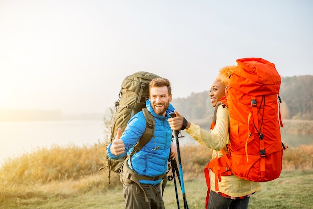 Porträt eines schönen multiethnischen Paares, das im Herbst mit Rucksäcken und Stöcken auf dem Campingplatz in der Nähe des Zeltes auf dem grünen Rasen wandert