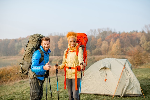 Porträt eines schönen multiethnischen Paares, das im Herbst mit Rucksäcken und Stöcken auf dem Campingplatz in der Nähe des Zeltes auf dem grünen Rasen wandert