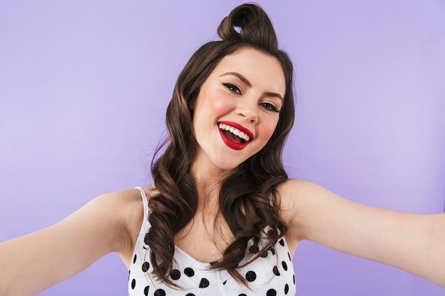 Porträt eines schönen Pin-up-Mädchens mit hellem Make-up, das isoliert über einer violetten Wand steht und ein Selfie macht