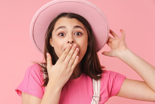 Porträt eines schönen schockierten Mädchens mit Hut, das Überraschung ausdrückt und die Kamera einzeln über rosafarbener Wand betrachtet