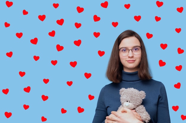 Porträt eines schönen Studentenmädchens mit Brille, das ein graues Teddybärspielzeug auf blauem Hintergrund mit roten Herzen umarmt