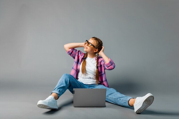 Porträt eines Schulmädchens, das Laptop benutzt, während es mit gekreuzten Beinen sitzt, isoliert auf grauem Hintergrund