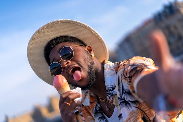 Porträt eines schwarzen ethnischen Mannes, der den Sommerurlaub am Strand genießt, ein Eis isst und ein Selfie macht