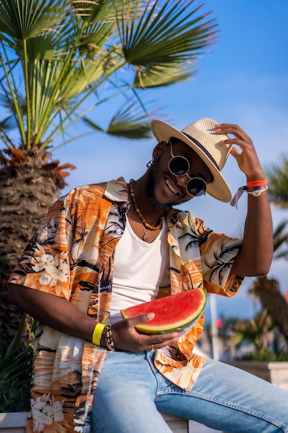 Porträt eines schwarzen ethnischen Mannes, der mit Hut und Wassermelone den Sommerurlaub am Strand genießt