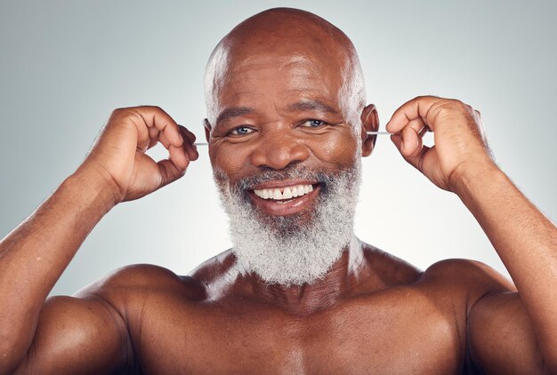 Porträt eines schwarzen Mannes, der das Ohr mit einem Lächeln putzt Körperpflege und -pflege isoliert auf grauem Hintergrund Morgenroutine Gesundheit und glücklicher älterer Mann mit Ohrstöpseln in den Ohren, um für Wellness im Studio zu reinigen