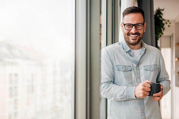 Porträt eines selbstbewusst lächelnden Geschäftsmannes, der am Fenster im Büro Kaffee genießt