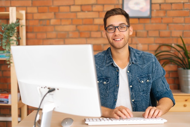 Porträt eines selbstbewussten, fröhlichen, gutaussehenden jungen, freiberuflichen Designers mit stylischer Brille, der an einem Desktop-Computer arbeitet, der am Schreibtisch zu Hause sitzt