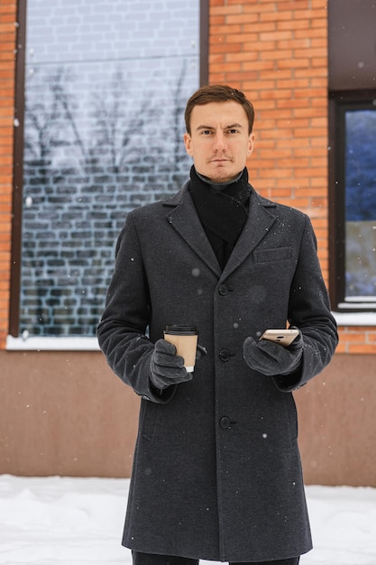 Porträt eines selbstbewussten Geschäftsmannes im Wintermantel mit einer Tasse Kaffee mit Smartphone in der Stadtstraße