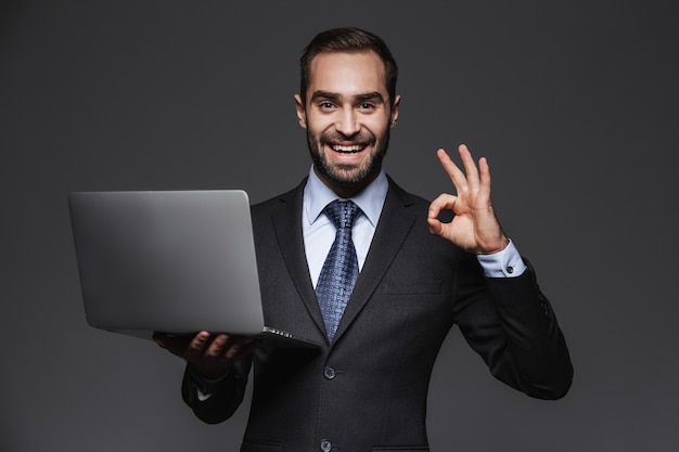 Porträt eines selbstbewussten gutaussehenden Geschäftsmannes, der Anzug isoliert trägt und Laptop-Computer hält, ok